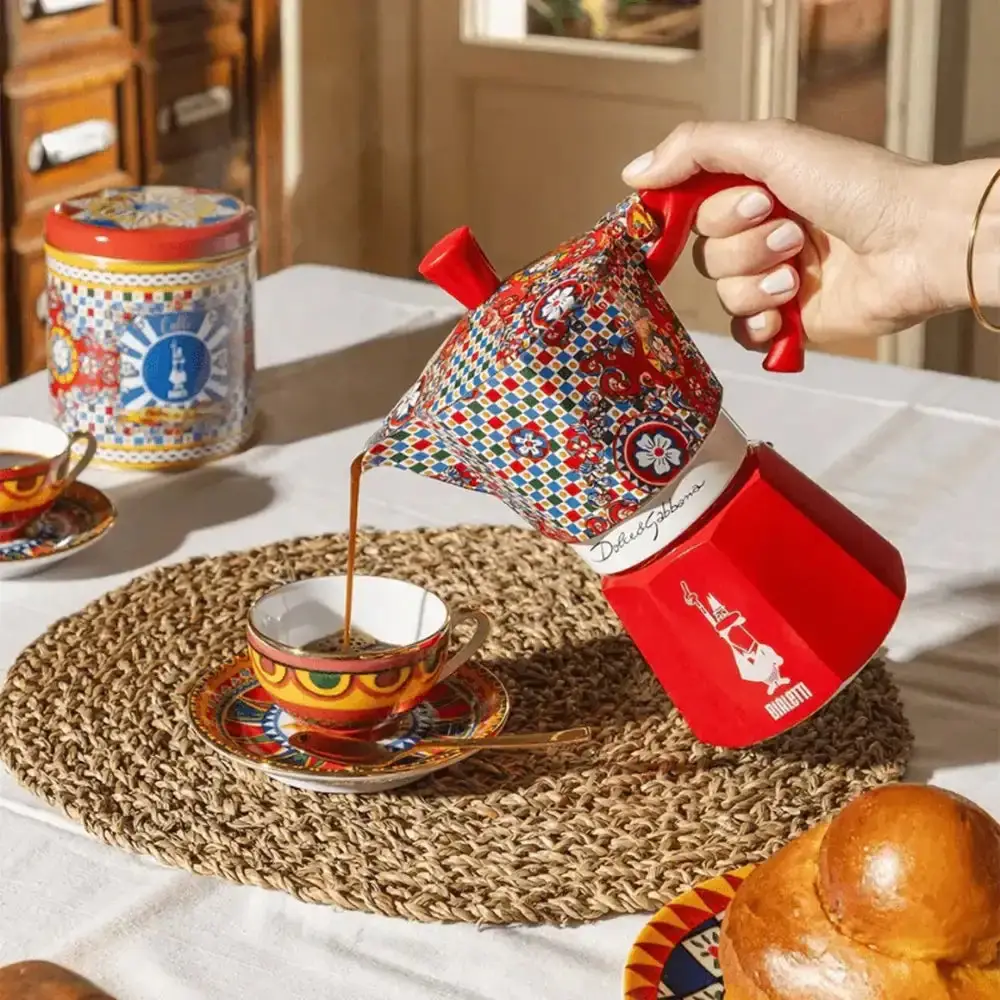 A brightly patterned Bialetti stovetop moka pot pouring espresso into a decorative cup