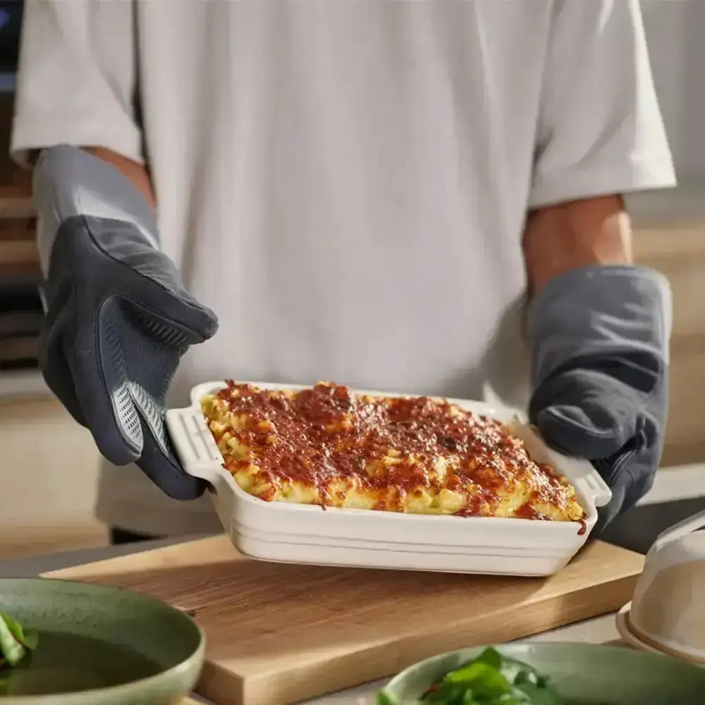 A person wearing grey oven gloves carrying a hot baking dish from the oven