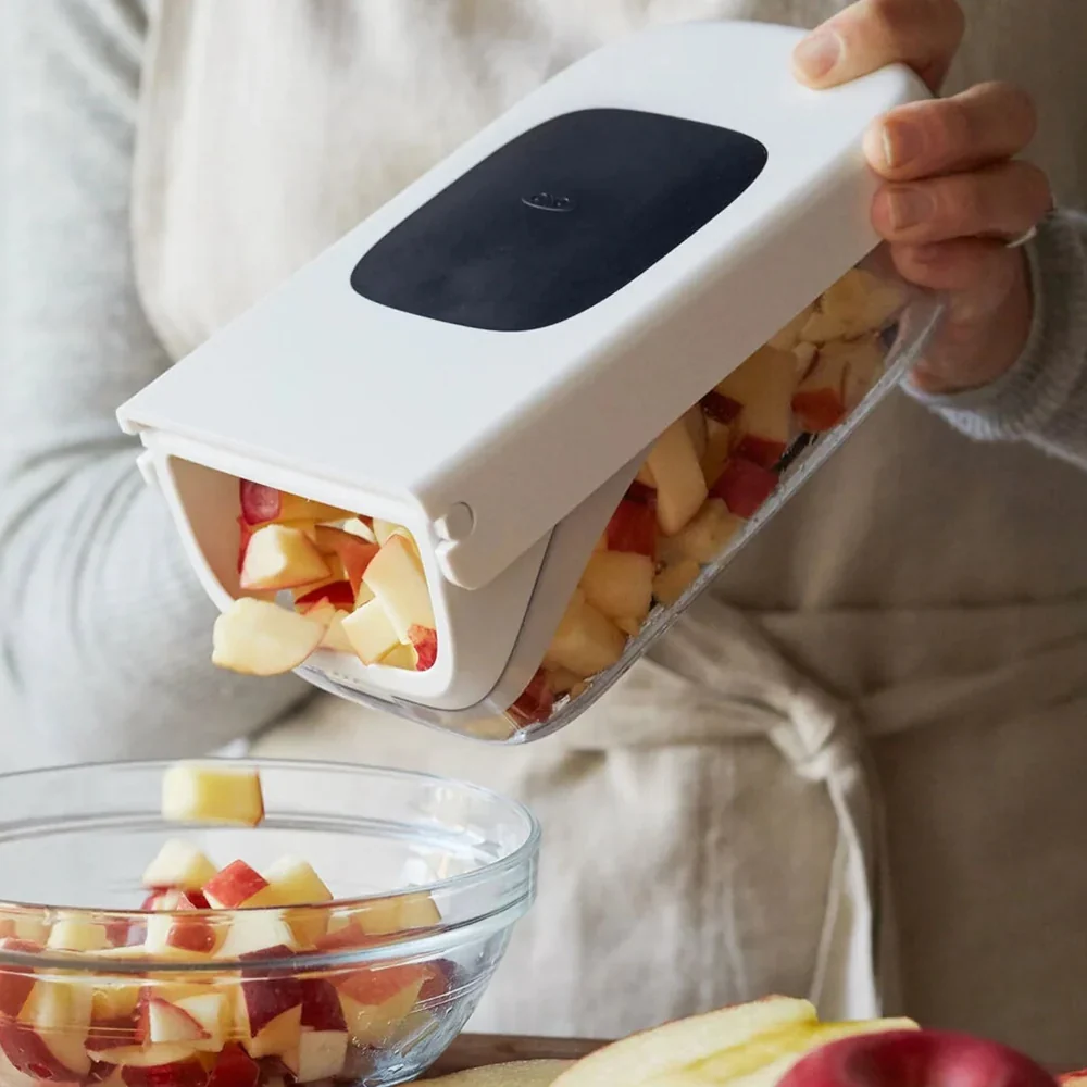 Hands pouring chopped apple from a white vegetable chopper into a glass bowl