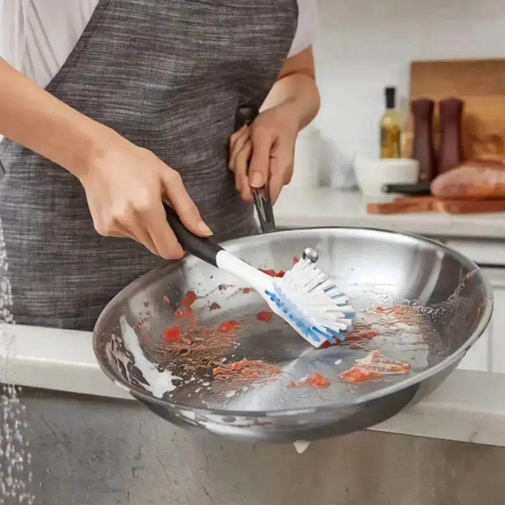 A hand scrubbing a stainless steel frying pan with a blue and white washing-up brush over a sink