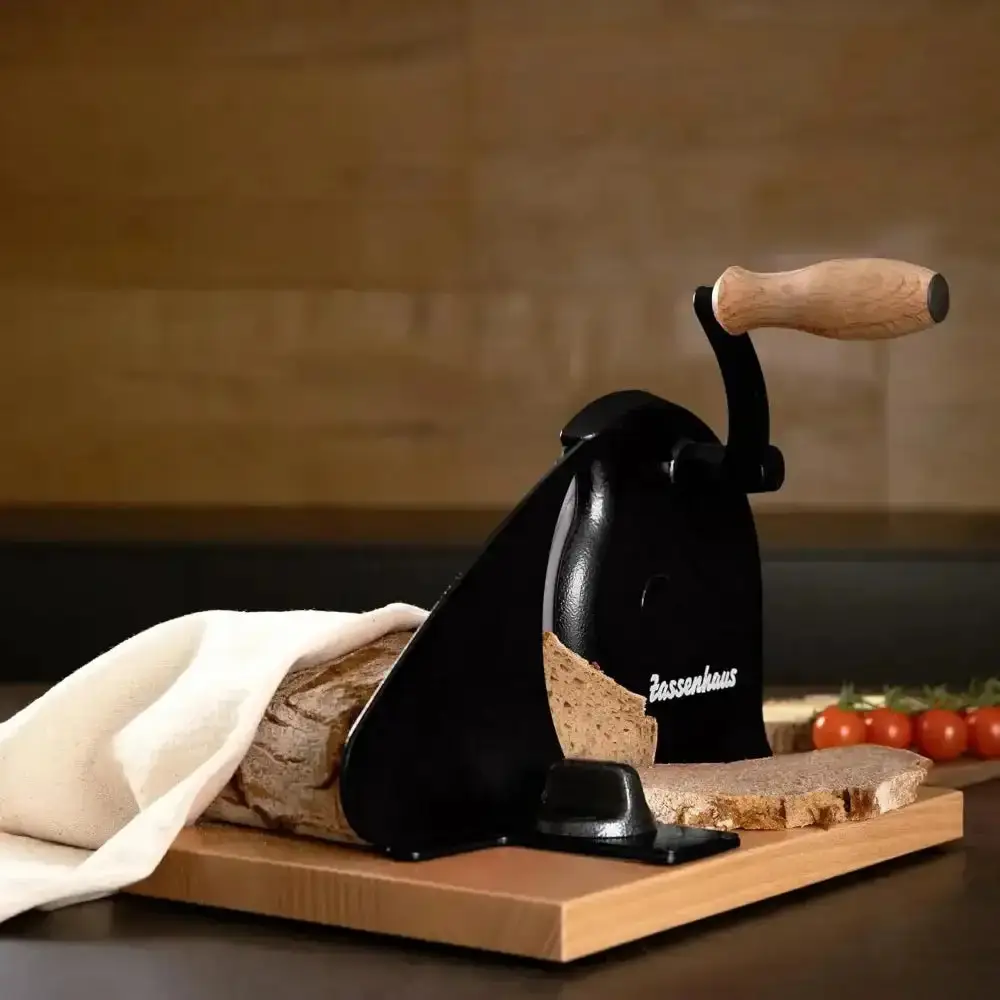 A black hand-crank bread slicer cutting a rustic loaf on a wooden board