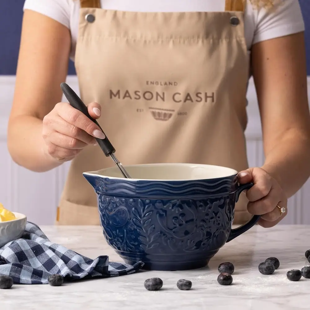 A baker whisking batter in an embossed navy Mason Cash mixing bowl with blueberries on a marble counter