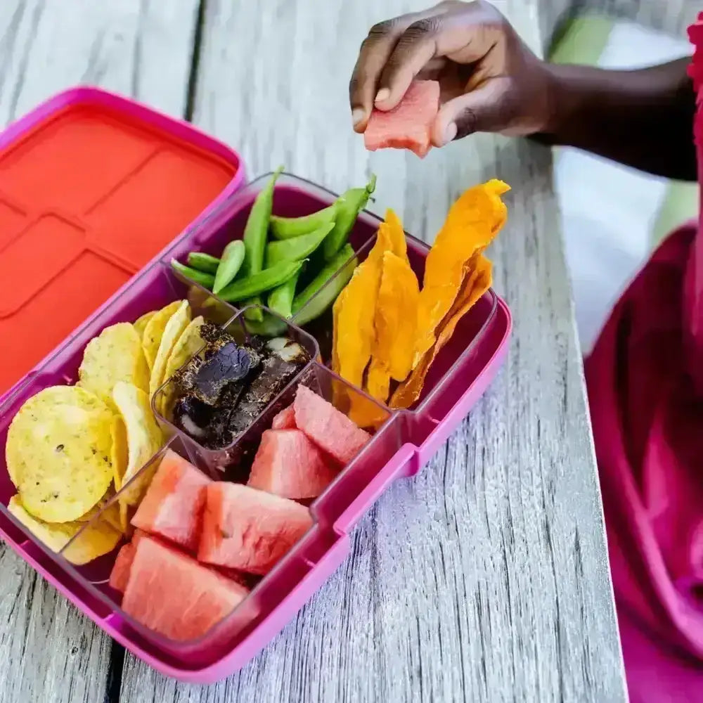 A child packing watermelon, snap peas and snacks into a pink Crunchbox lunch box