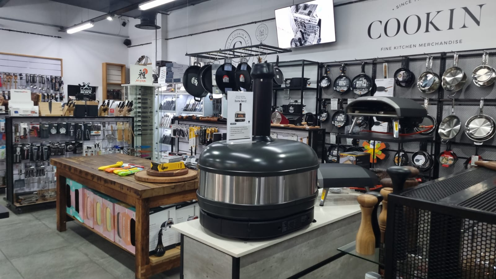 Inside Cookin: pizza ovens, hanging pans, knives and kitchen tools under the Cookin Fine Kitchen Merchandise sign
