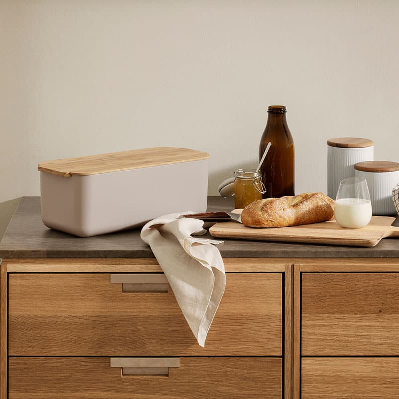 Cream bread bin with a wooden lid on a kitchen counter beside a crusty baguette on a wooden board