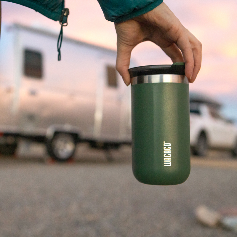 A green Wacaco insulated travel tumbler held in a hand outdoors at sunset