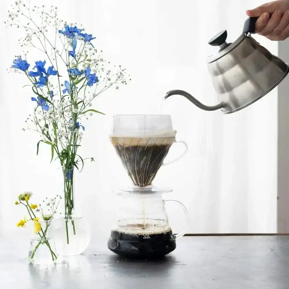 A stainless gooseneck kettle pouring hot water over a Hario V60 dripper set on a glass coffee server