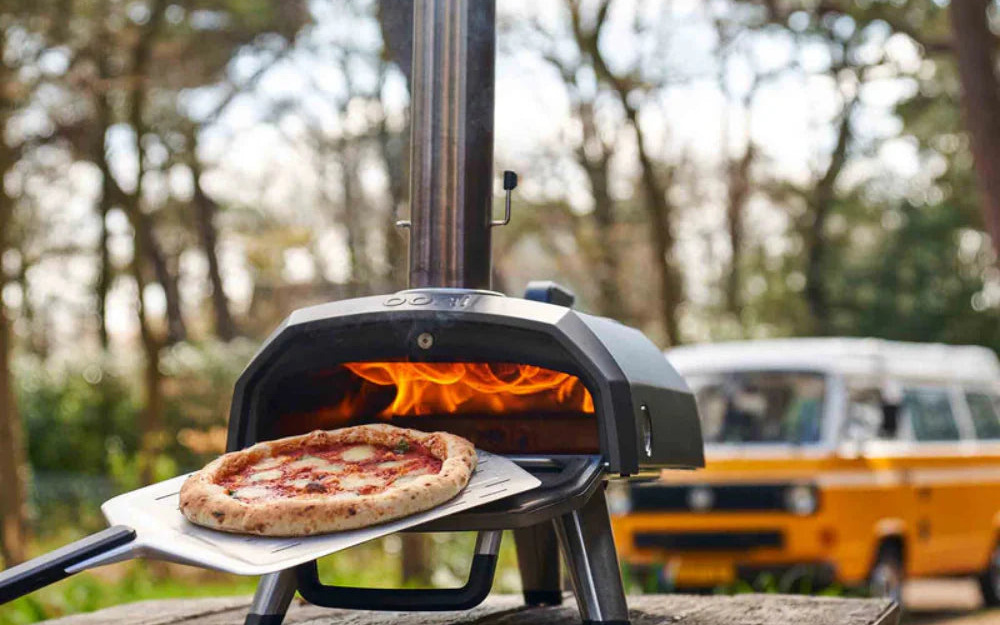 A lit Ooni Karu 2 outdoor pizza oven with a pizza on the peel in a wooded campsite