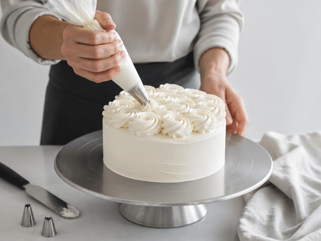 Hands piping buttercream rosettes onto a cake on a metal turntable beside a set of piping nozzles
