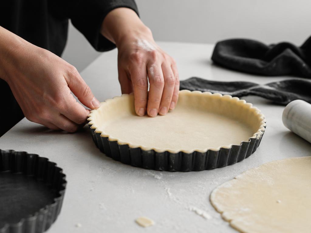 Hands pressing shortcrust pastry into a fluted loose-base quiche tin beside a baked pie and quiche