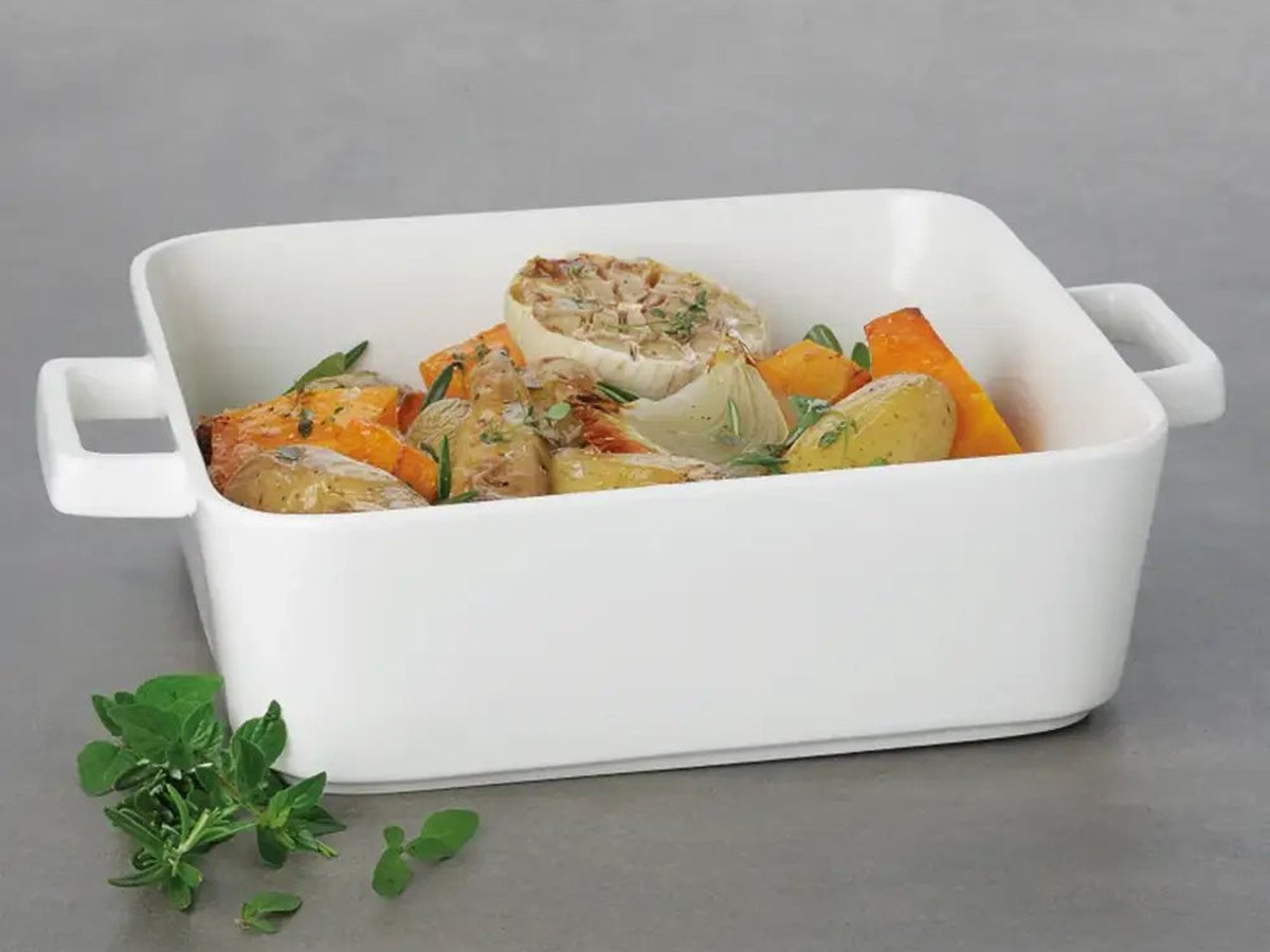 A white stoneware square baker of roasted vegetables and fish with fresh herbs on a pale grey surface