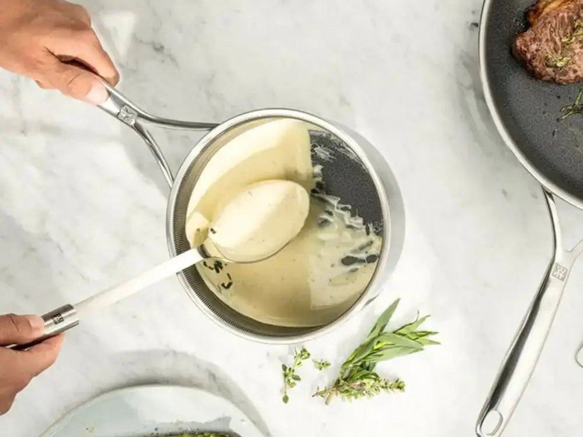 Hollandaise being ladled from a stainless steel saucepan beside asparagus on a pale marble counter
