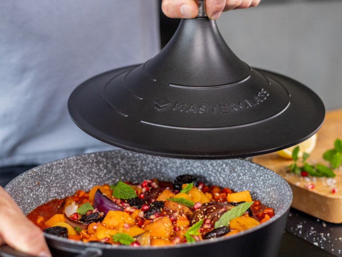 A hand lifting the conical lid from a cast-aluminium tagine of bubbling vegetable stew on a timber board