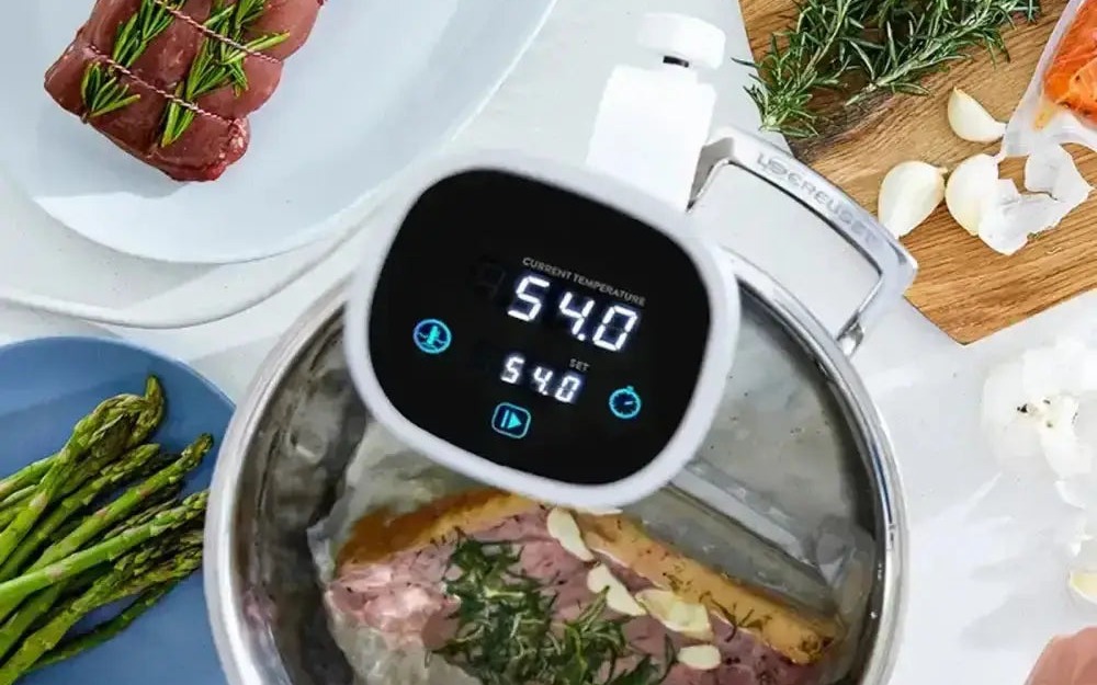 A sous-vide circulator holding a water bath with steak, herbs and vegetables alongside