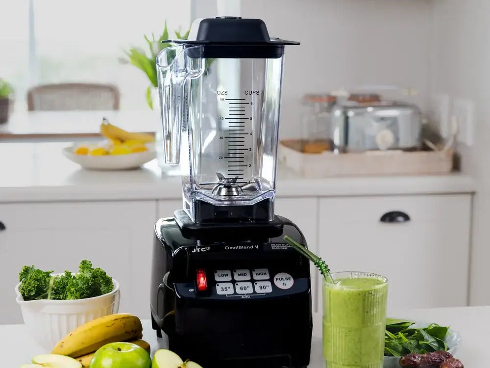 An OmniKitchen Omniblend V jug blender on a bright kitchen counter with fruit and a green smoothie