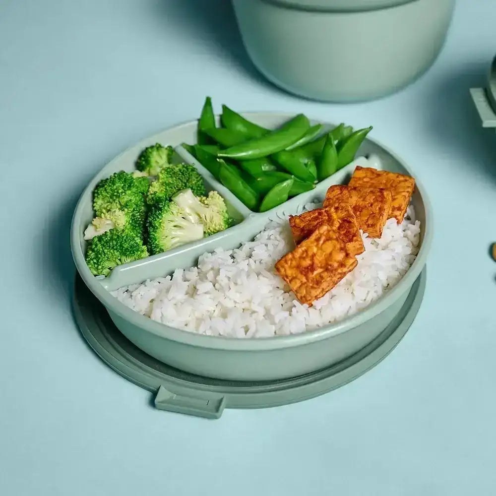 A Mepal Cirqula bento bowl in sage holding rice, broccoli and sugar snap peas on a pale blue table