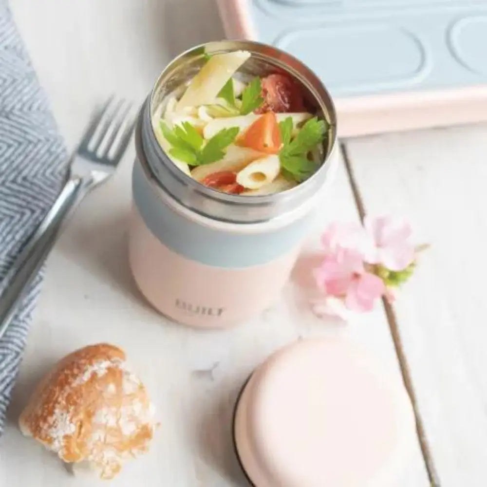 A pale pink Built insulated food flask filled with pasta salad on a white table with a fork and a bread roll