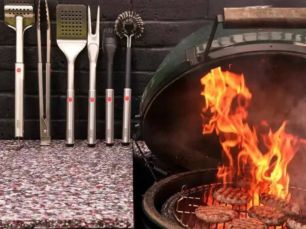 Stainless steel braai tools lined up against a brick wall beside a kettle braai with flames and burgers grilling